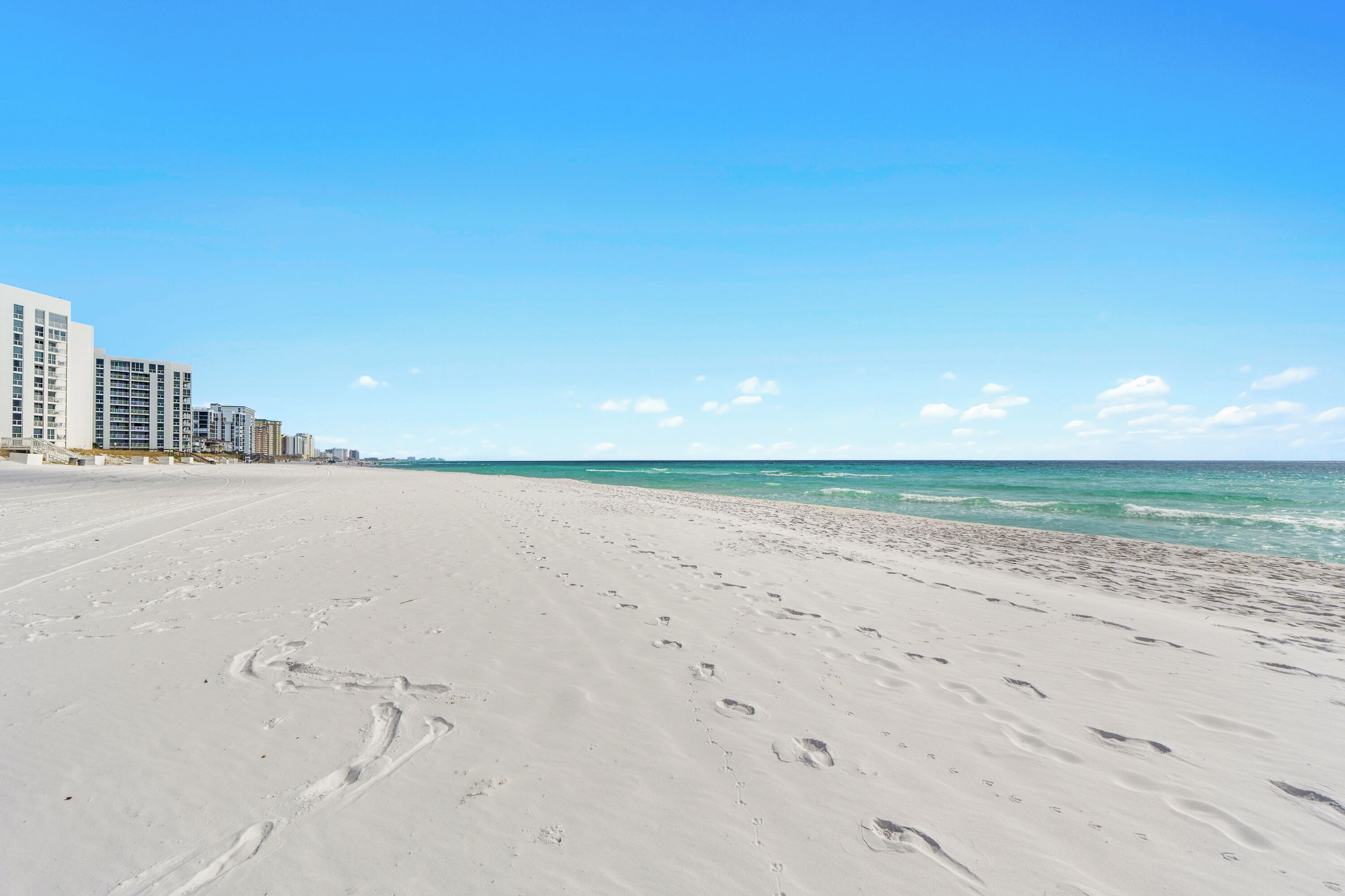 Une vaste plage de sable parsemée d’empreintes, un ciel bleu dégagé et des bâtiments visibles à l’arrière-plan.
