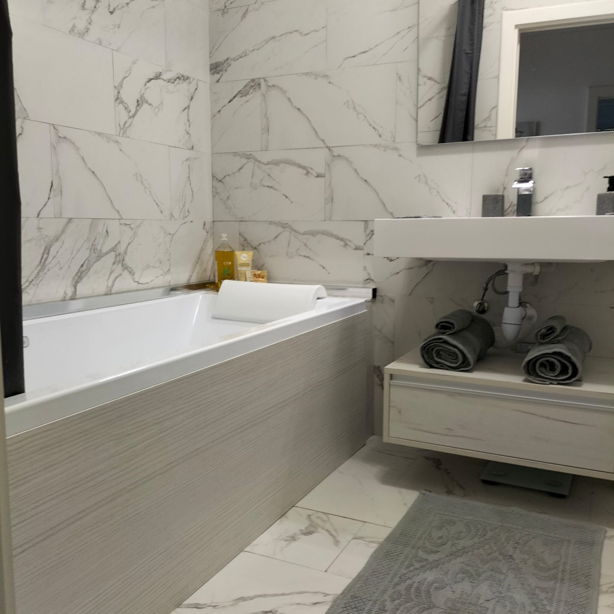A bathroom with a white marble wall, a white bathtub, a white sink with a mirror above it, and a small shelf with towels and toiletries.