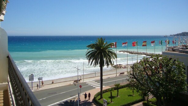 Vista costera desde un balcón con vista a una playa, palmeras, una carretera y banderas.