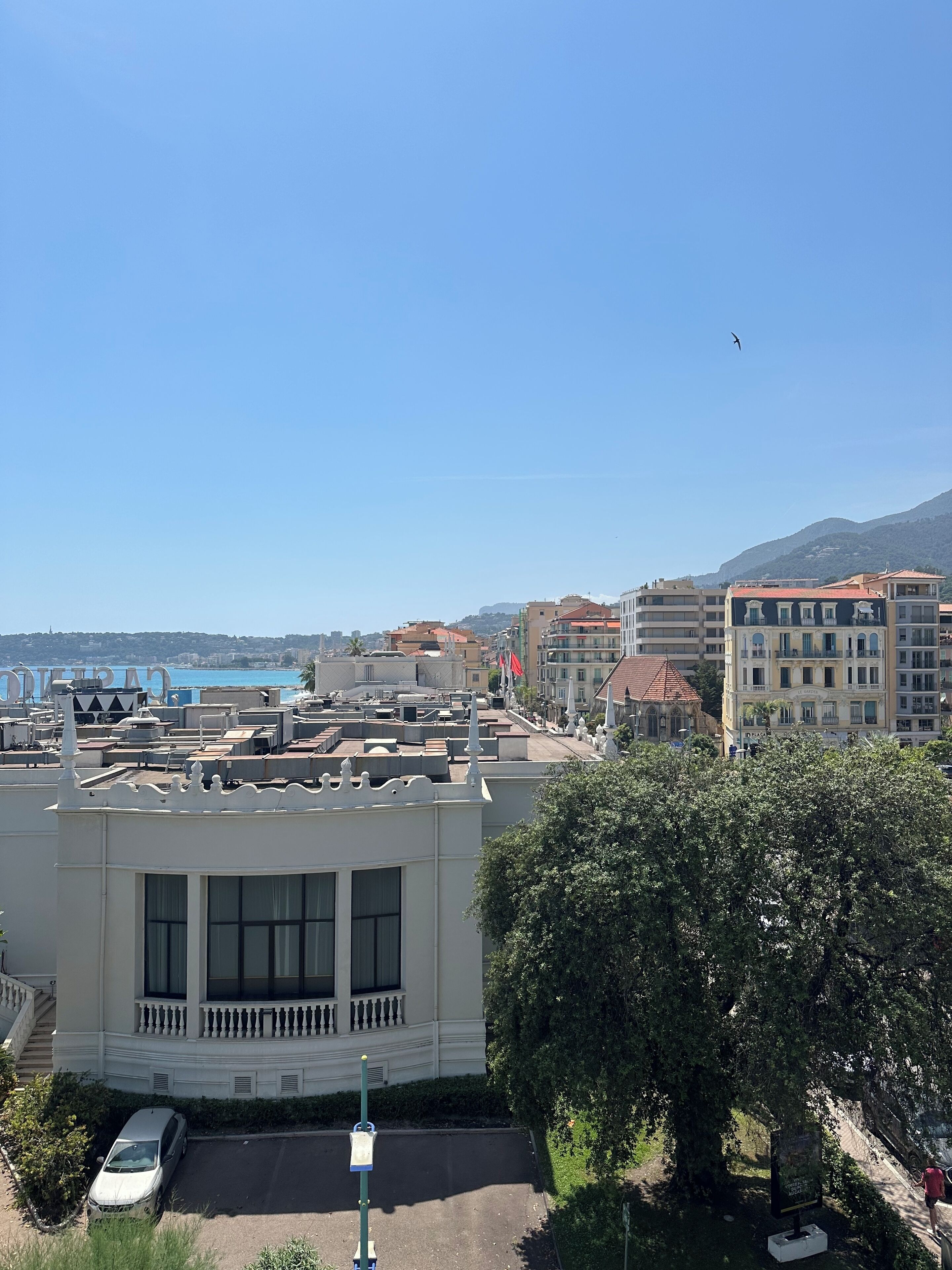 A coastal cityscape with a prominent building featuring large windows and a balcony, surrounded by other buildings and a clear sky.