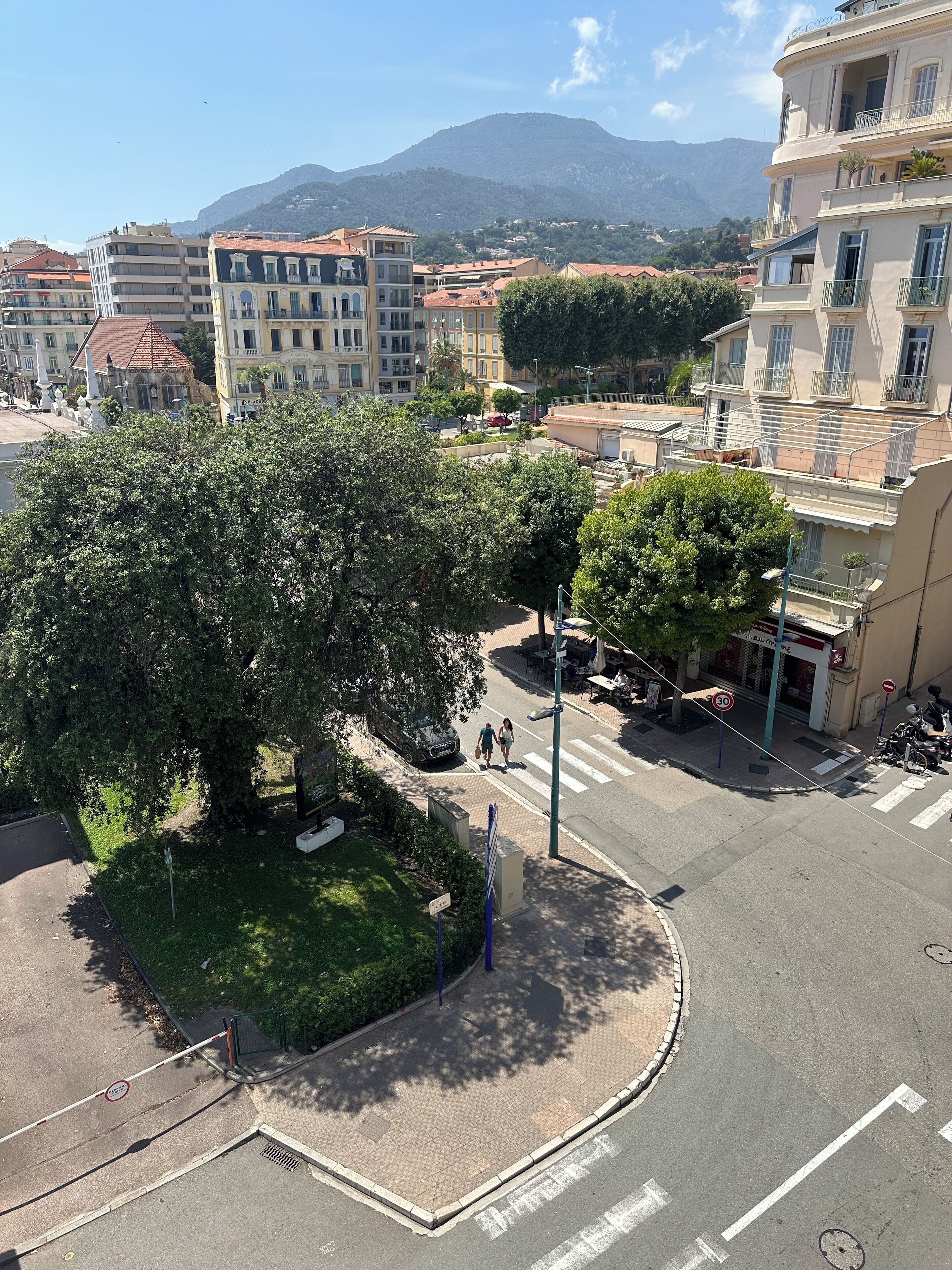 A roundabout with a central grassy area, surrounded by buildings with balconies and a mountain in the background.