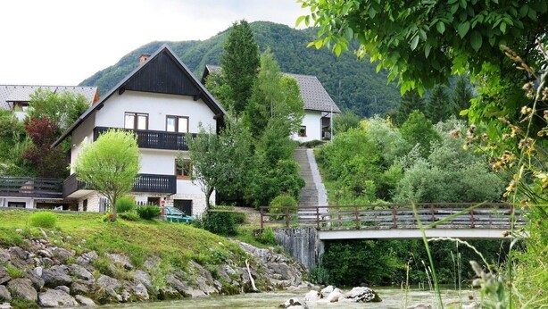 Un edificio blanco con techo negro y balcón, rodeado de vegetación y un río.