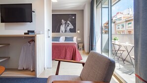 Une chambre d’hôtel avec un lit, une chaise, une télévision, un tableau et un balcon donnant sur des immeubles.