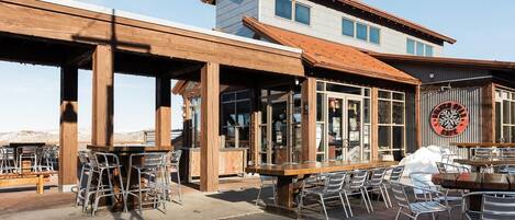 An outdoor patio with wooden tables and chairs, a covered seating area, and a building with large windows and a distinctive circular logo.