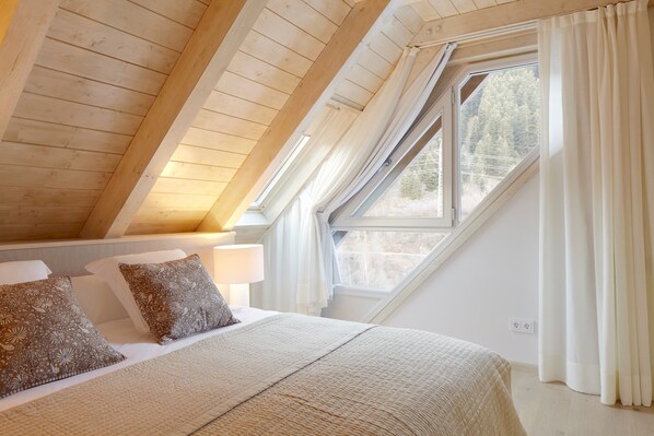A bedroom with a sloped ceiling, a large window with curtains, a bed with patterned pillows, and a bedside lamp.