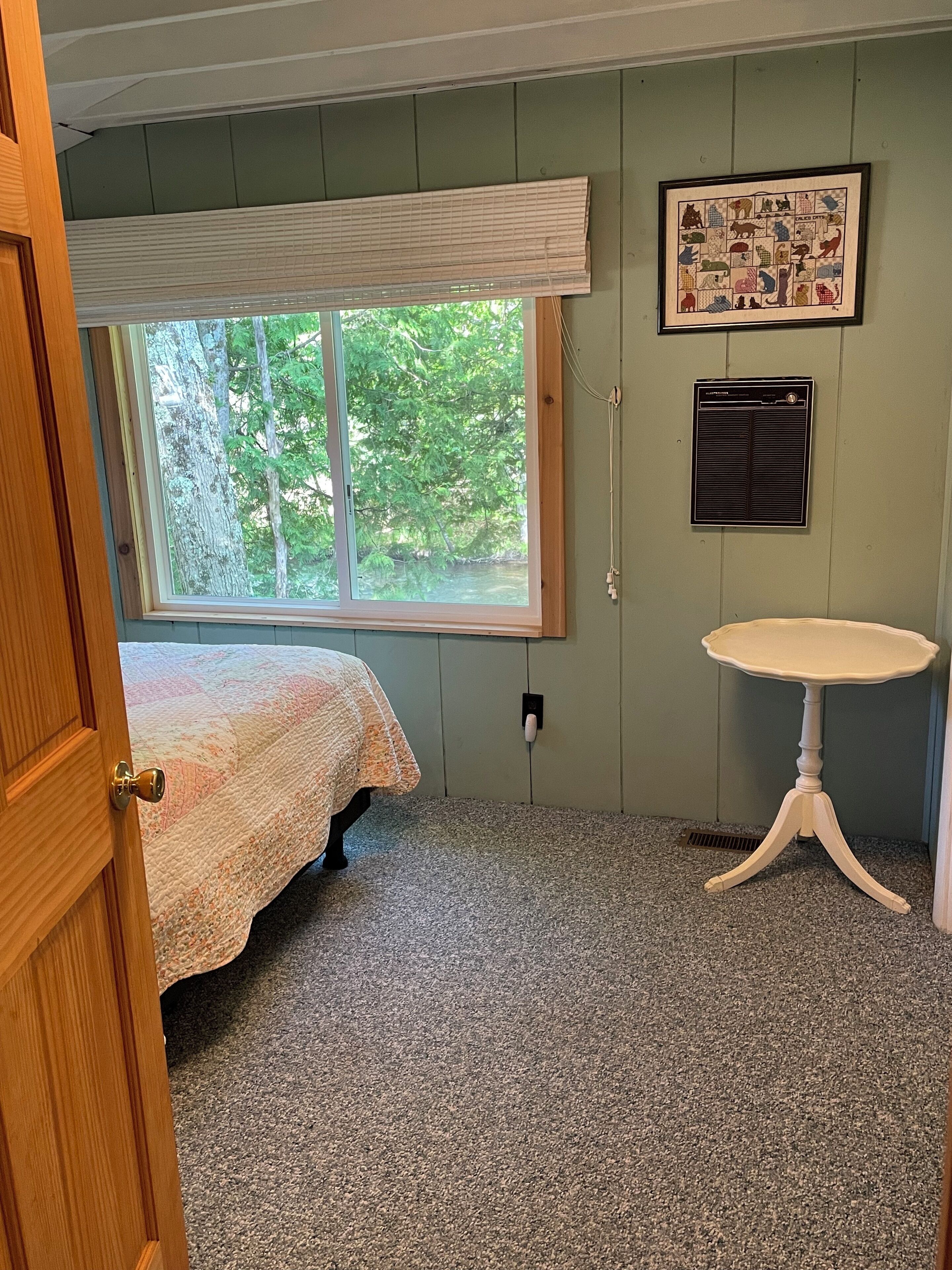 A bedroom with a bed, a window with a view of trees, a wall-mounted air conditioner, and a small table.