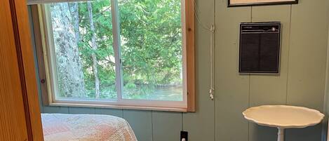 A bedroom with a bed, a window with a view of trees, a wall-mounted air conditioner, and a small table.