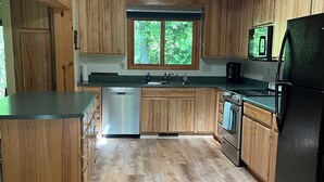 A kitchen with wooden cabinets, a black refrigerator, and a window with greenery outside. - Relaxing cozy cabin on beautiful Pere Marquette River - Recently Renovated!! (Branch)