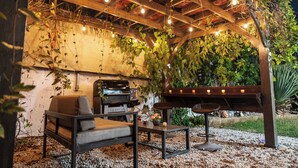 An outdoor patio with a wooden pergola, string lights, a grill, a cushioned chair, and a table with candles. - Private Patio Retreat with Zen Vibe and Laundry (Ponce)