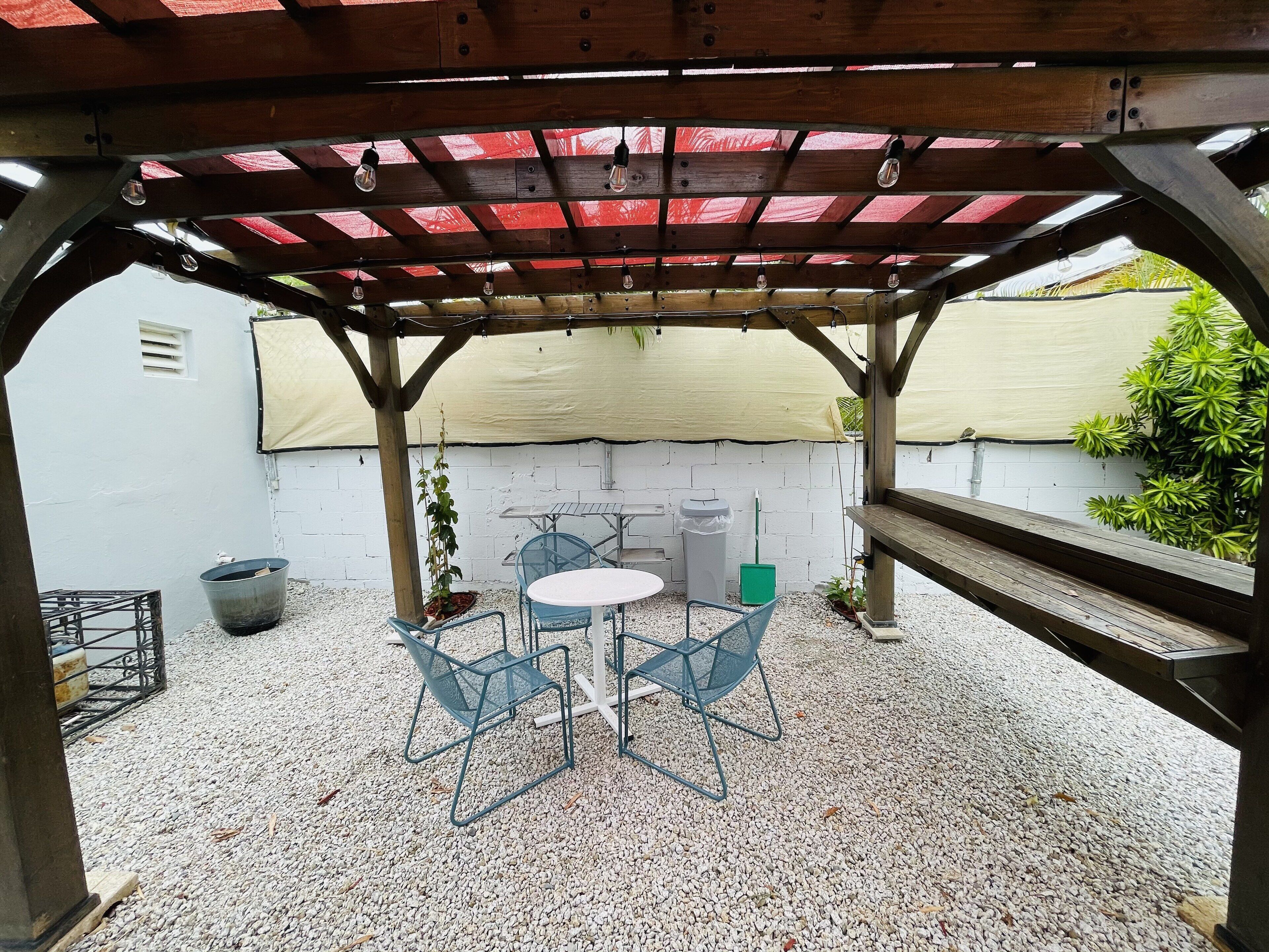 Een overdekt terras met een tafel, stoelen, een afvalbak en een potplant.