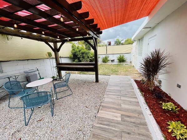 A covered patio area with a table and chairs, a wooden deck, and a garden bed with plants.