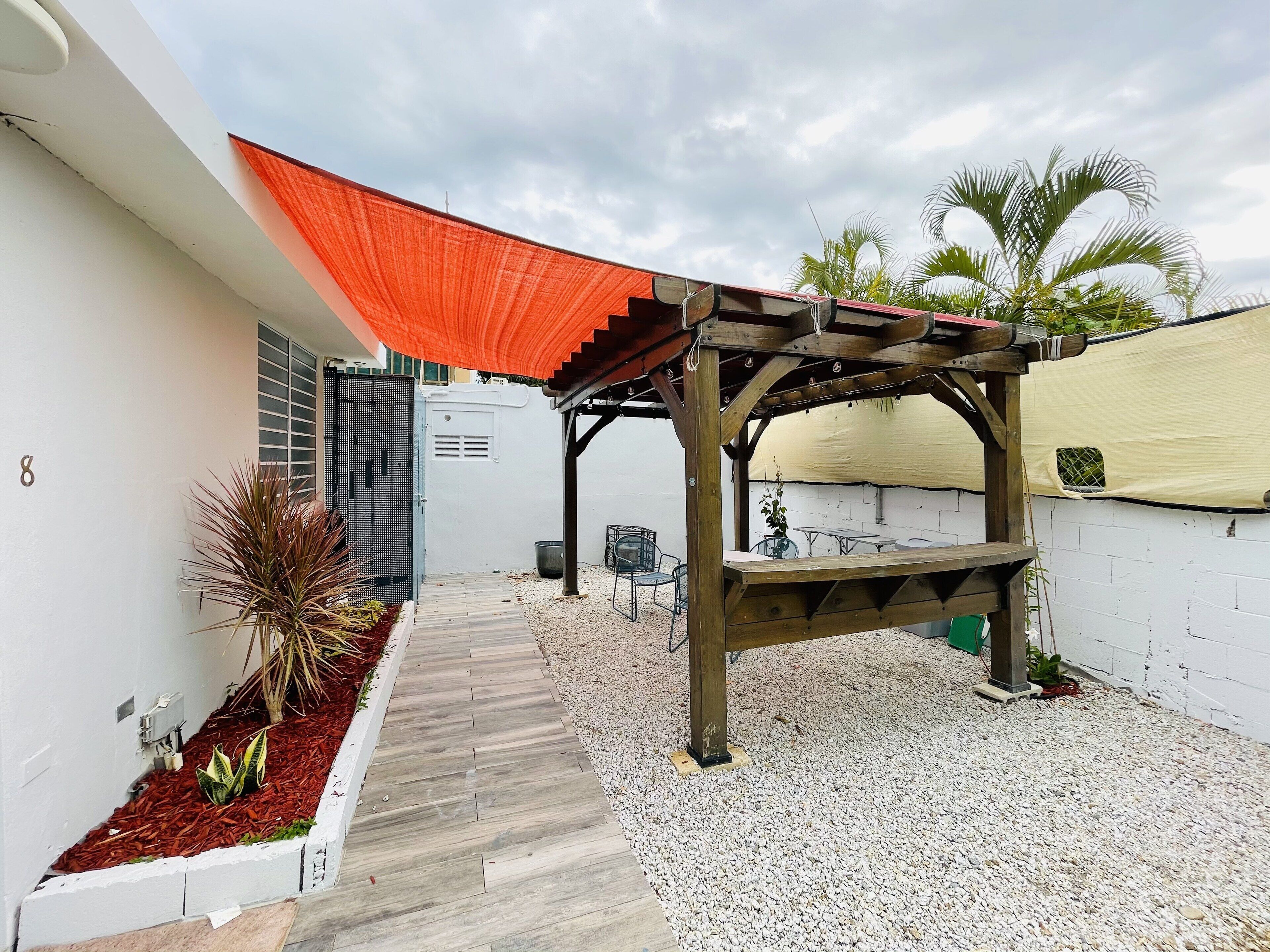 Un espace extérieur aménagé, abrité et doté d’une pergola en bois, d’un chemin de gravier et d’un mur blanc orné du chiffre « 8 ».