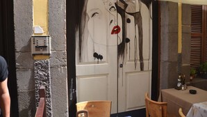 Une porte ornée d’une fresque représentant le visage d’une femme aux lèvres rouges, une petite table avec des bouteilles et une chaise.