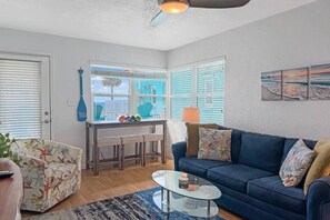 A living room with a sofa, a glass coffee table, a patterned armchair, a dining table with chairs, and a view of the outside.