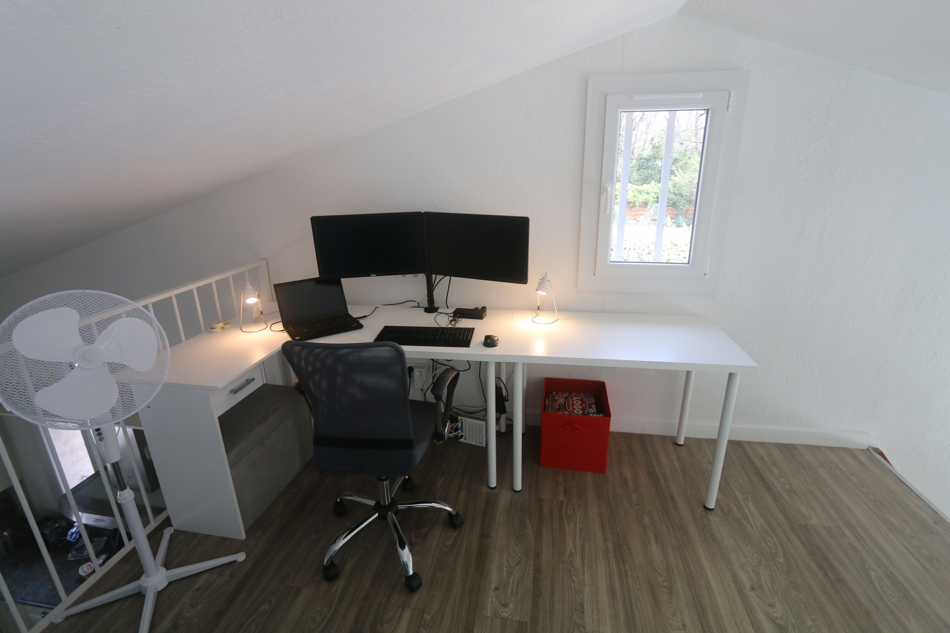 Een klein wit bureau met een computer en beeldscherm in een kamer met houten vloer en een raam.