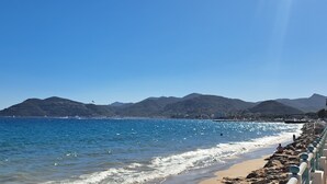 Une plage à l’eau d’un bleu cristallin, avec un rivage rocheux et des montagnes en arrière-plan.