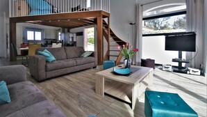 Un vaste espace de vie comprenant un canapé, une table basse et un téléviseur. Un escalier en bois mène à une mezzanine.