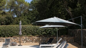 Un espace aménagé au bord de la piscine, avec des chaises longues et des parasols, entouré de murs en pierre et d’arbres.