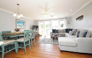 A spacious living room with a grey sofa, teal chairs, a wooden dining table, and a ceiling fan.