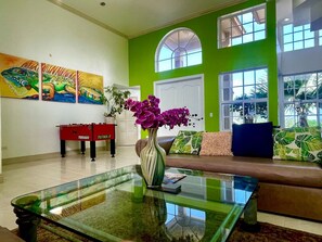 A modern living room with a glass coffee table, a sofa with patterned cushions, a red pool table, and a large window with a view of greenery.