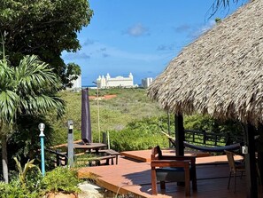 A wooden deck with outdoor seating, a thatched-roof structure, and a view of a large building in the distance.