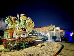 Una propiedad residencial bien iluminada de noche, con entrada de coches, un vehículo estacionado y un jardín ajardinado con palmeras y flores rojas.