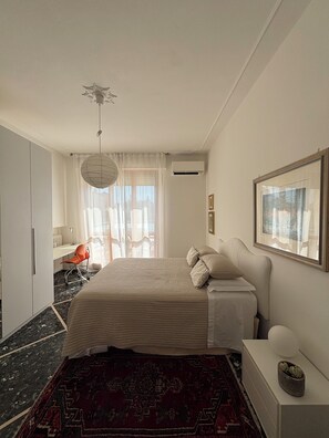 Habitación de hotel con cama, escritorio, silla, un ventanal grande y un cuadro en la pared.