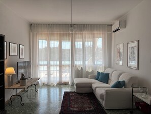 Un salon avec un canapé blanc, un tapis à motifs rouges, une lampe et des œuvres d’art accrochées aux murs.