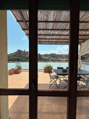 Vista desde una habitación con puerta de madera que da a una terraza exterior con mesa y sillas, plantas en macetas y una vista despejada de un cuerpo de agua y edificios al fondo.
