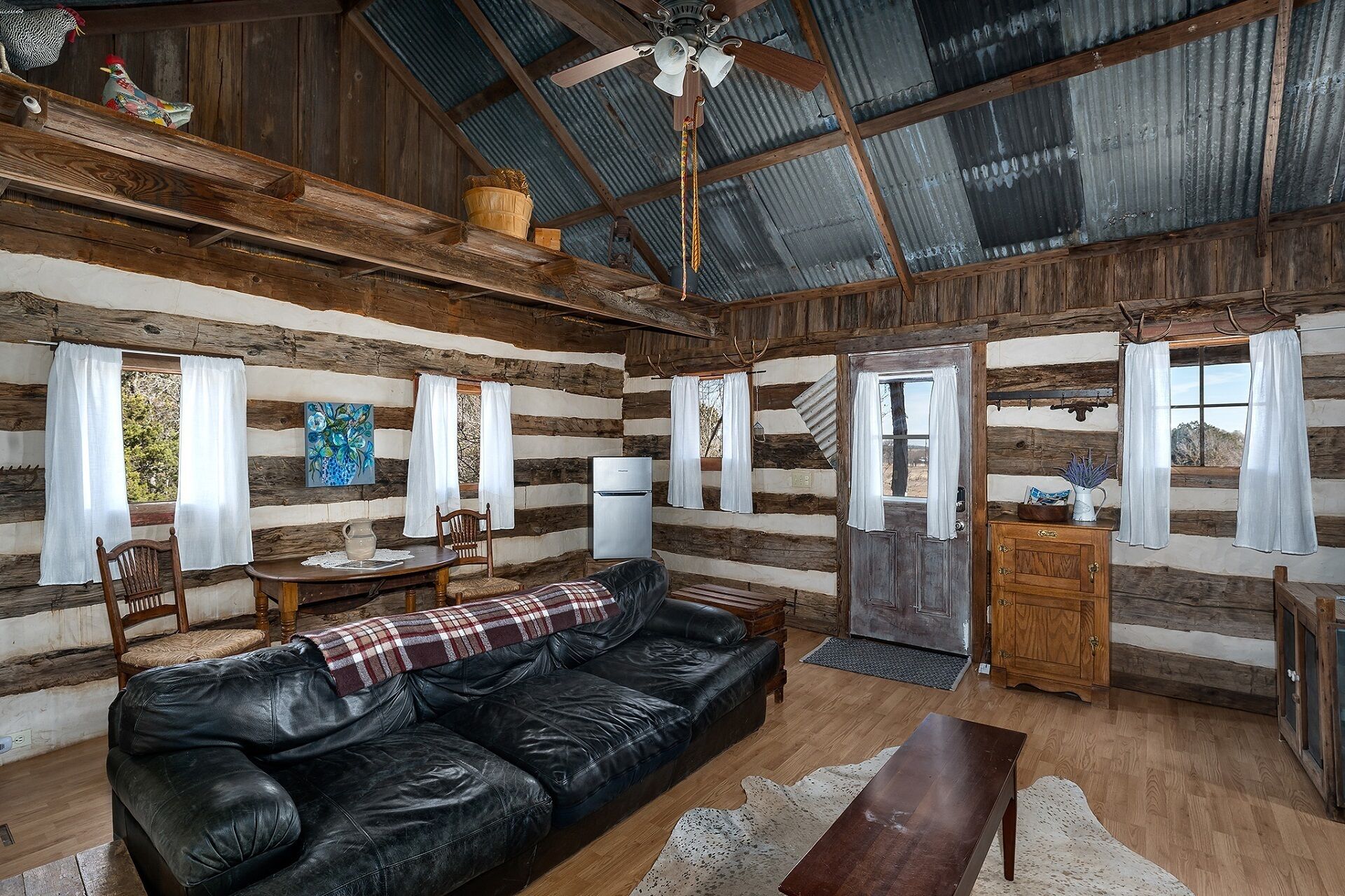 A rustic cabin interior with a leather sofa, wooden dining table, and a ceiling fan.