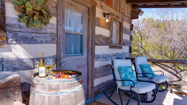 Une cabane rustique avec un tonneau en bois, une bouteille de vin et une assiette de fruits sur le porche. On y trouve deux fauteuils à bascule avec des coussins.