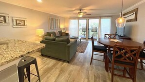 A modern living room with a green sofa, a dining table with chairs, a ceiling fan, and a large window with a view of the outdoors. - Pool&Golf Views 2BR Retreat with FreeBeachService (Destin)