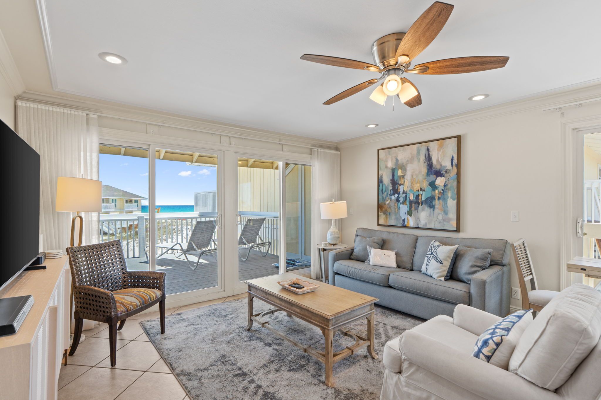 A living room with a television, seating, a ceiling fan, and a view of the beach.