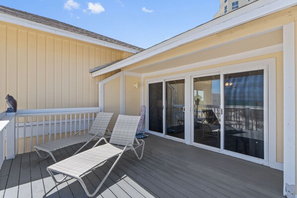 A covered outdoor deck with two lounge chairs and a table, a sliding glass door leading to an indoor area, and a railing along the deck's edge. - Beachside Escape | Spacious 1BR with Balcony (Destin)