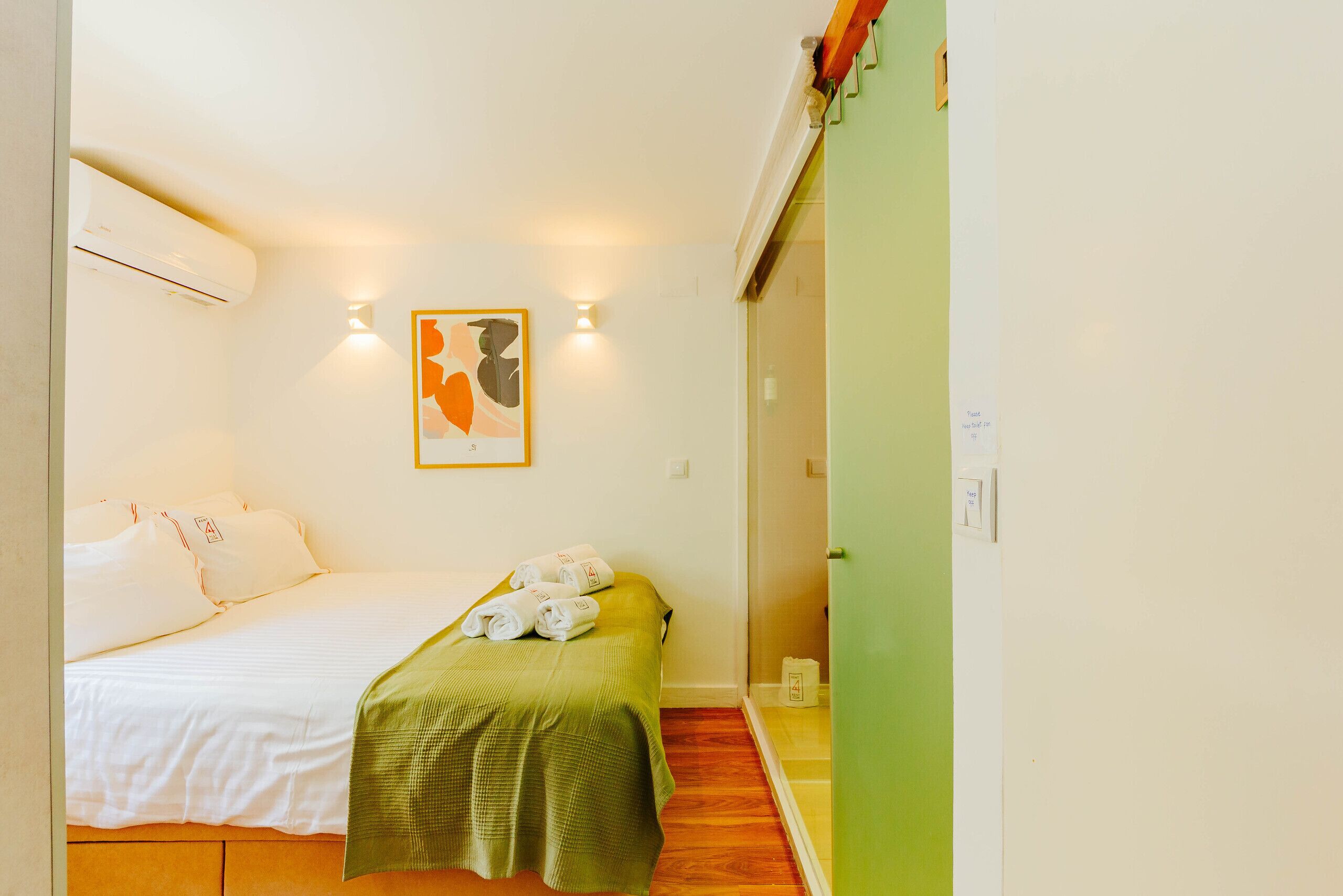 Une petite pièce propre, avec un lit, une couverture verte et une œuvre d’art accrochée au mur.