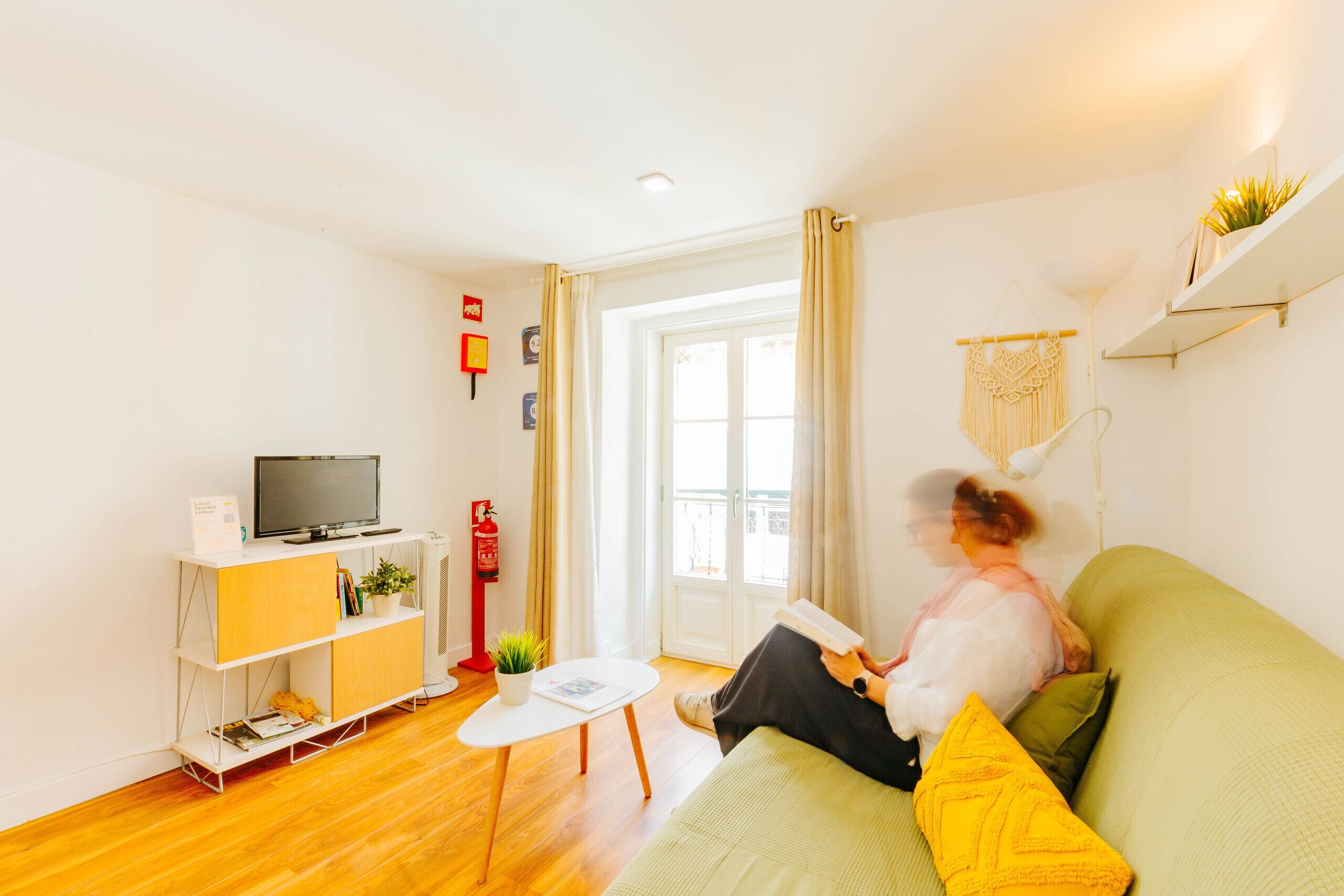 A living room with a green sofa, a white coffee table, a TV on a wooden cabinet, and a window with curtains.