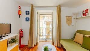 A compact living space with a sofa, a television, a small table with a plant, a fire extinguisher, and a door leading to an outdoor balcony. - RENT4REST BAIRRO ALTO CHARMING 1 BEDROOM APARTMENT (Lisboa ciudad)