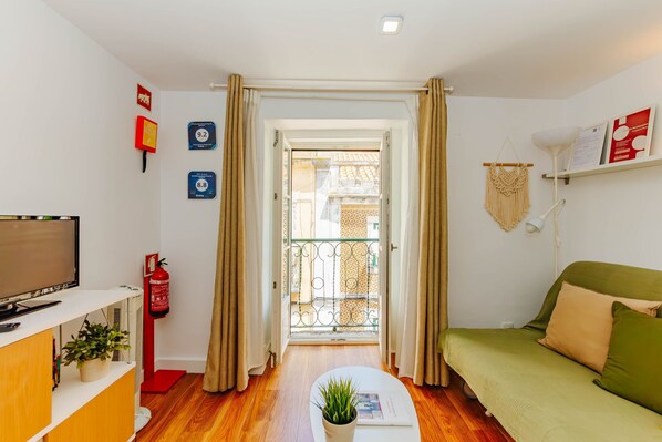 A compact living space with a sofa, a television, a small table with a plant, a fire extinguisher, and a door leading to an outdoor balcony. - RENT4REST BAIRRO ALTO CHARMING 1 BEDROOM APARTMENT (Lisboa ciudad)
