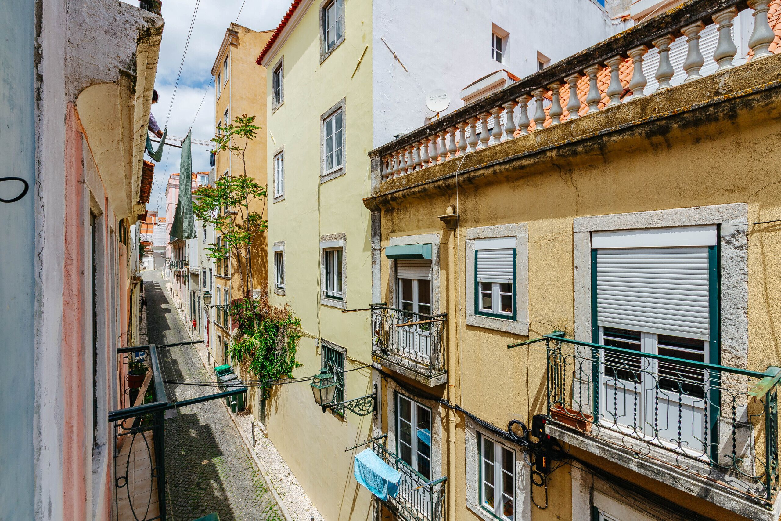 A narrow street with buildings on either side, featuring balconies with railings and windows with shutters.