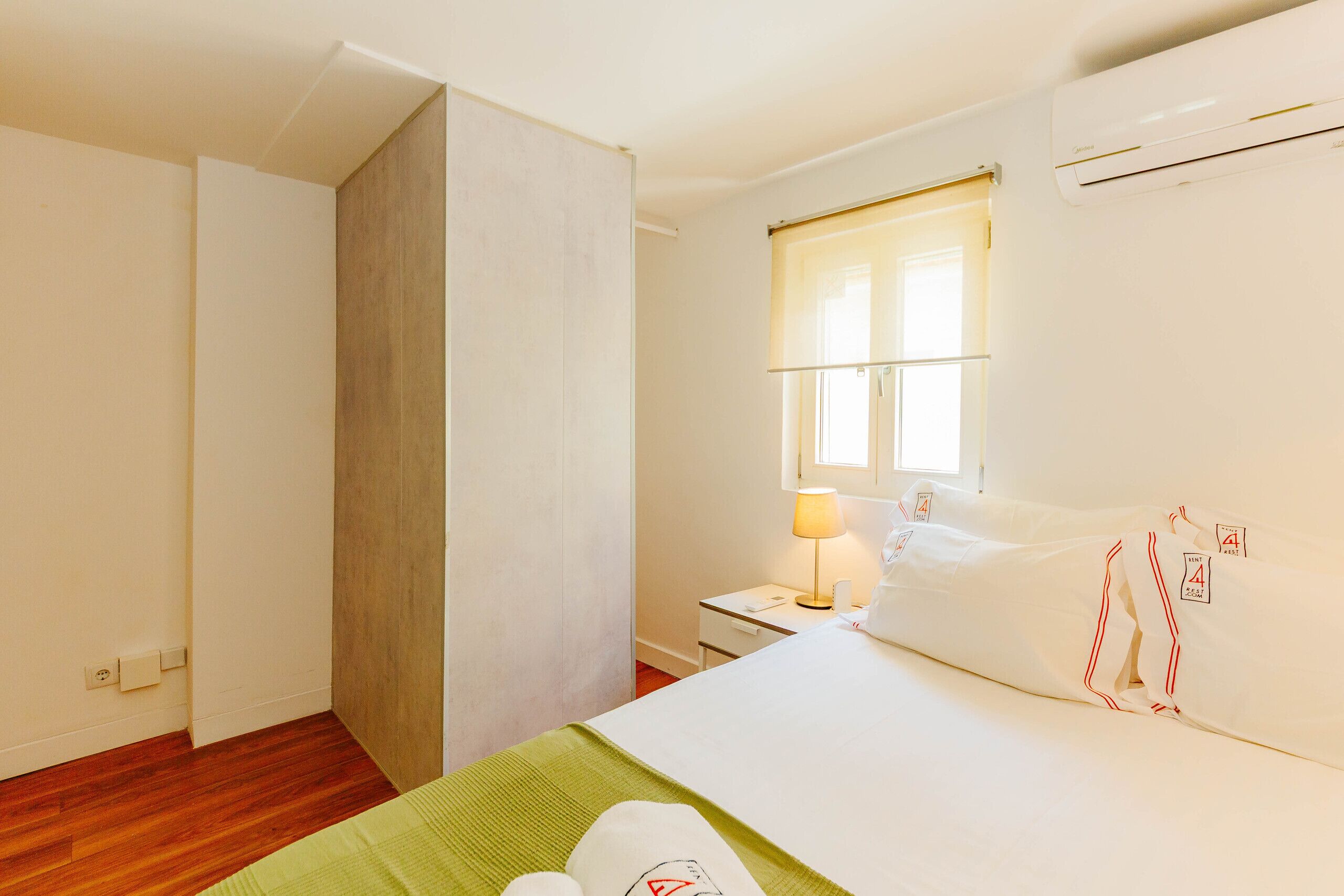 A hotel room with a bed, a nightstand, a lamp, a window with blinds, and an air conditioning unit.