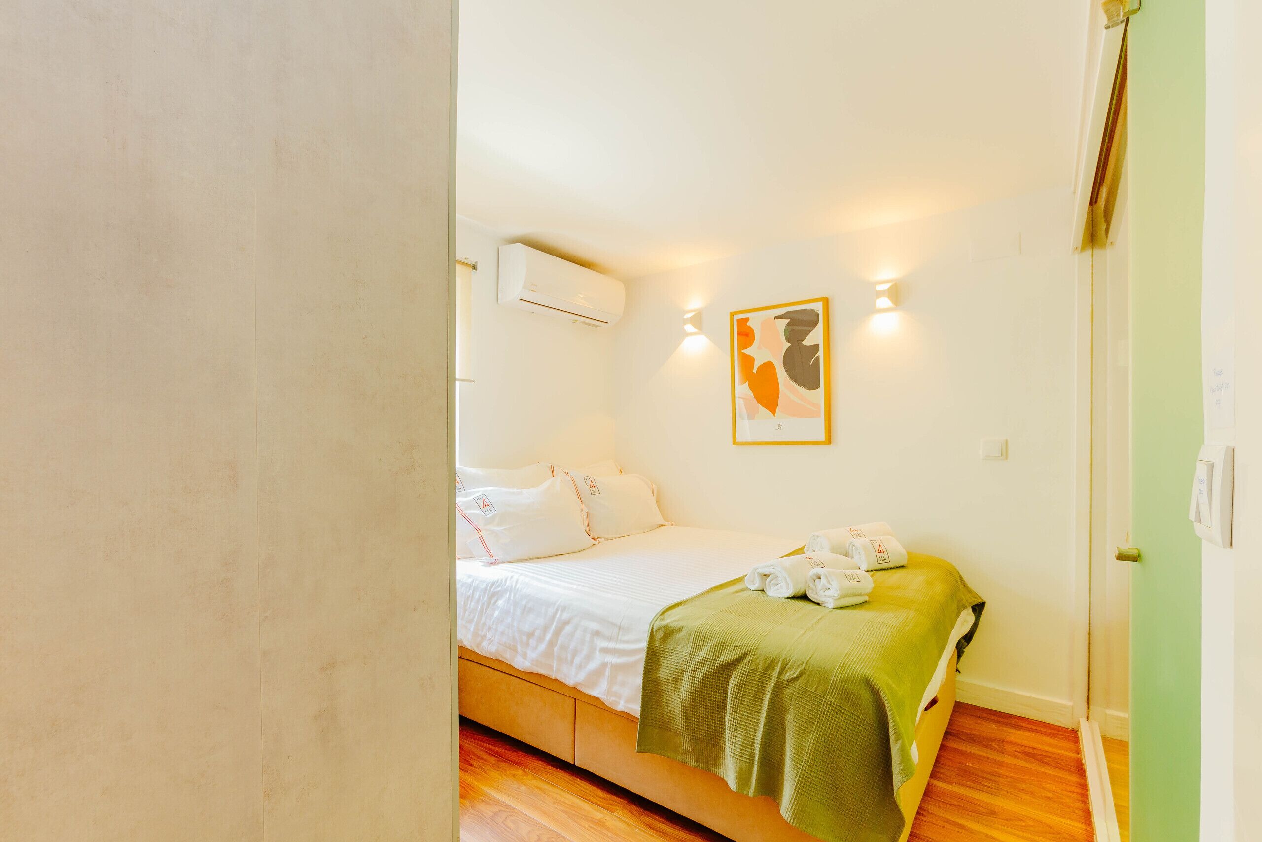 A bedroom with a bed, pillows, a green blanket, and a wall-mounted air conditioner.