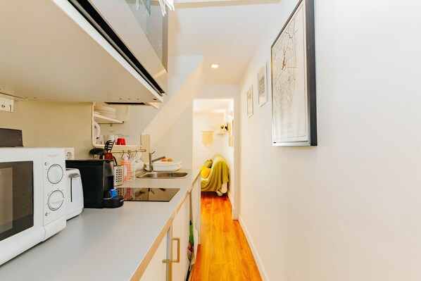 A narrow hallway with a kitchen area on the left, featuring a microwave and sink. A person is seen in the distance, and there are framed pictures on the wall. - RENT4REST BAIRRO ALTO CHARMING 1 BEDROOM APARTMENT (Lisboa ciudad)