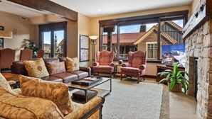 A spacious living room with a brown leather sofa, patterned armchairs, and a coffee table. Large windows offer views of a mountain landscape.