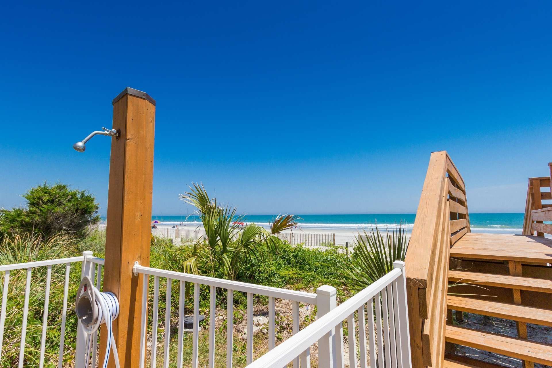 Een houten terras met een witte reling, met uitzicht op een strand met helderblauw water en een zandstrand.
