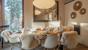 A dining area with a wooden table set for six, surrounded by white chairs with fluffy accents. A large mirror hangs above the table, reflecting the room.