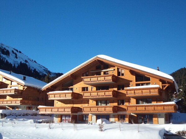 Un edificio de madera de varios pisos con balcones, rodeado de nieve y montañas al fondo.