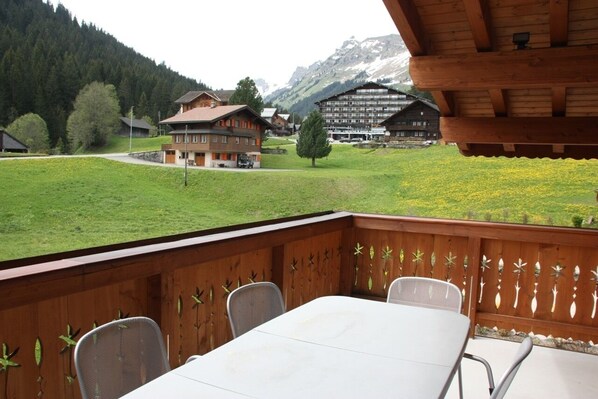 Eine Holzdielen-Terrasse mit Tisch und Stühlen, mit Blick auf ein grünes Feld und Gebäude in einer Berglandschaft.
