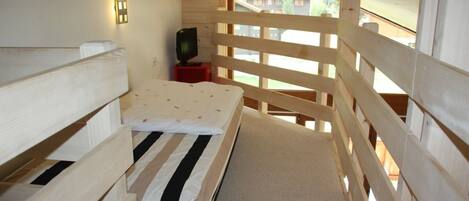 Habitación de cabaña de madera con literas, un televisor pequeño sobre un soporte rojo y una ventana con vistas a un área boscosa.