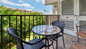 A balcony with a round table and two chairs, a door with louvered windows, and a view of a building and palm trees. - Open-Style Condo w/Chic Kitchen+Dining Area, WiFi, Lanai–Kamaole Sands 9206 (KIHEI)