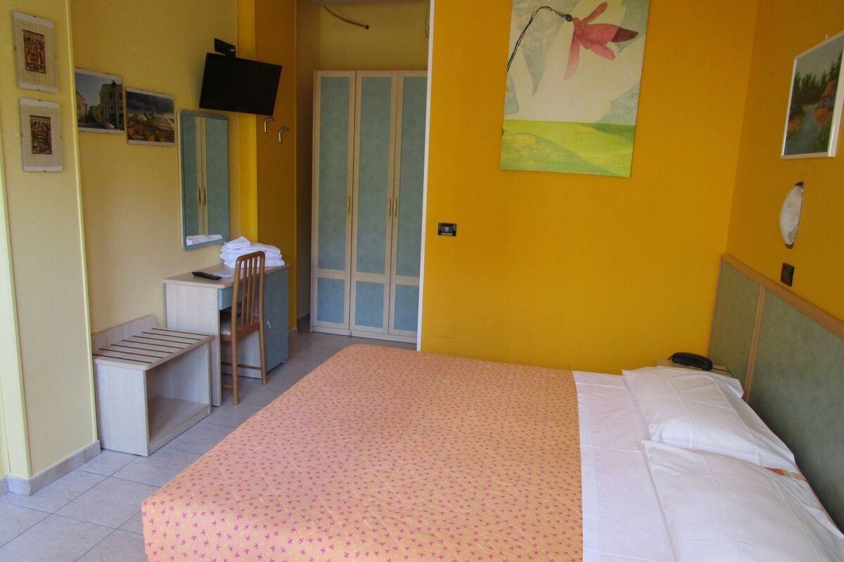 Quarto de hotel com cama, escrivaninha, cadeira, guarda-roupa e televisão na parede.